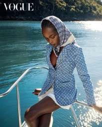 Model wearing a monogram print blue denim blazer and matching headscarf seated on a boat railing against open water