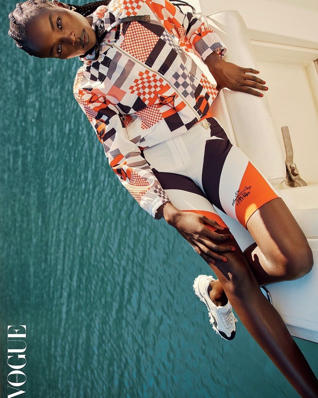 Fashion shot of a Black woman wearing a patterned jacket and shorts, posing on a boat with ocean water in the background and Vogue logo visible.