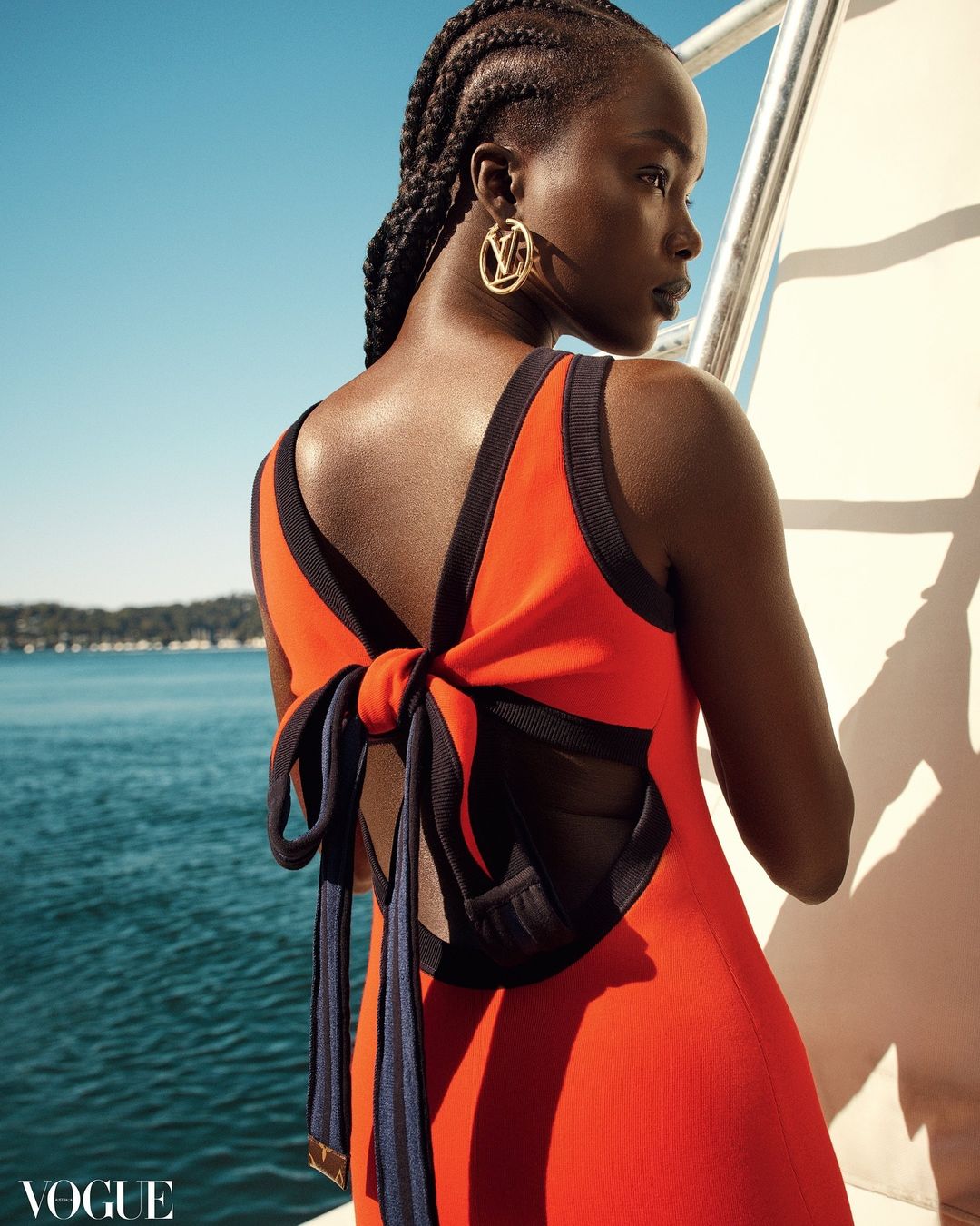 A woman with cornrow braids poses on a yacht, wearing a red dress with a large navy ribbon bow. Vogue logo is visible in the lower-left.