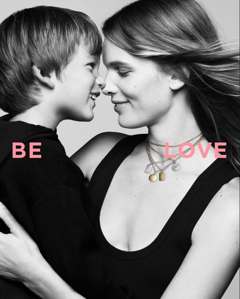 Mother and child touch noses in a black and white studio shot, the word 'LOVE' is visible in pink, with the models wearing layered necklaces.