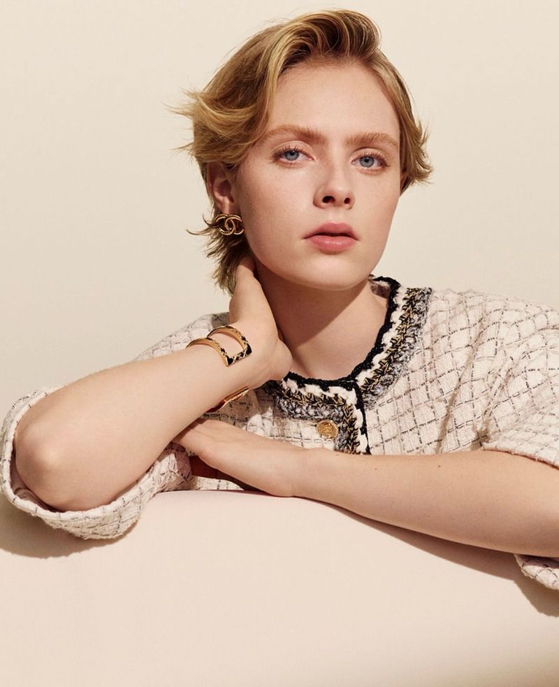 Blonde woman poses in Chanel jacket and gold jewelry against a neutral backdrop in a close-up studio fashion shot.