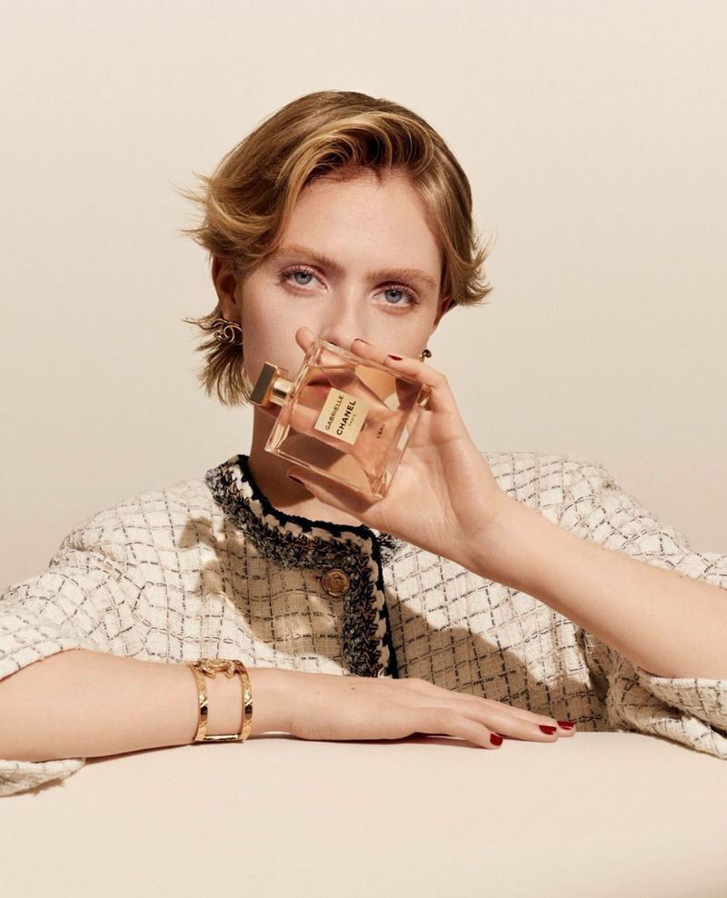 Adult woman poses indoors holding a Chanel perfume bottle. She wears tweed jacket, jewelry on neutral backdrop. Beauty advertising.
