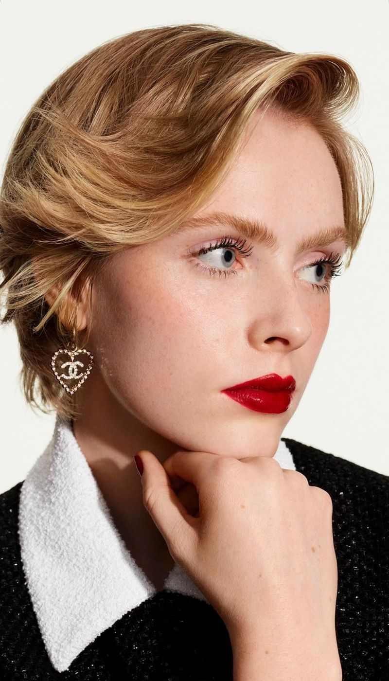Close-up shot of a fair-skinned adult female model with red lipstick, wearing a Chanel jacket and heart-shaped Chanel logo earrings.