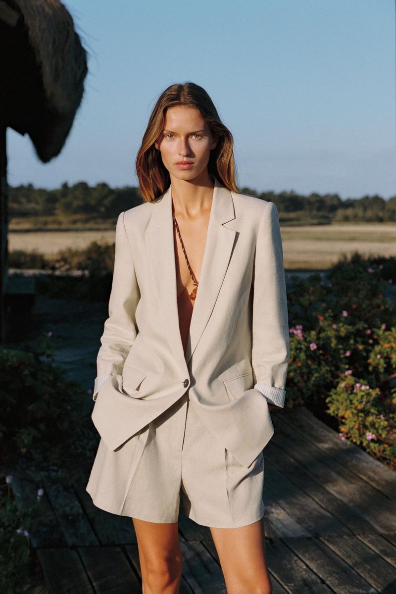 Adult female model in tan blazer and shorts posing outdoors, wearing a necklace, against a backdrop of foliage and a partly cloudy sky.