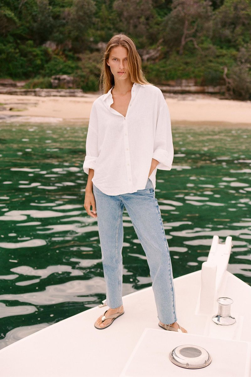 Woman in white blouse and jeans stands on white boat, sea and wooded coastline behind, natural lighting, medium shot, fashion.