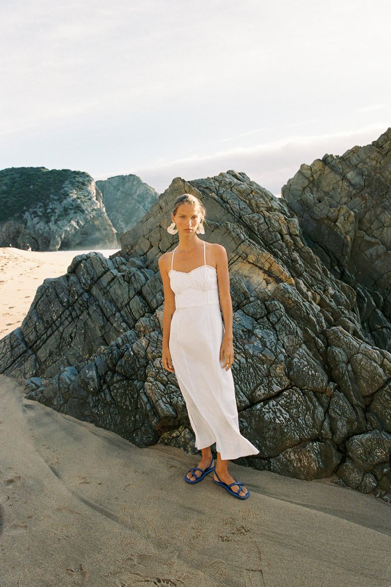 Adult woman in a white sundress poses on a sandy beach near rock formations on a sunny day, showcasing a fashion-forward outdoor look.