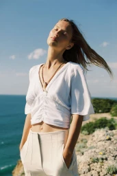 Model Lea Seelenmeyer wearing a white top and trousers with a necklace against an ocean and cliff background.