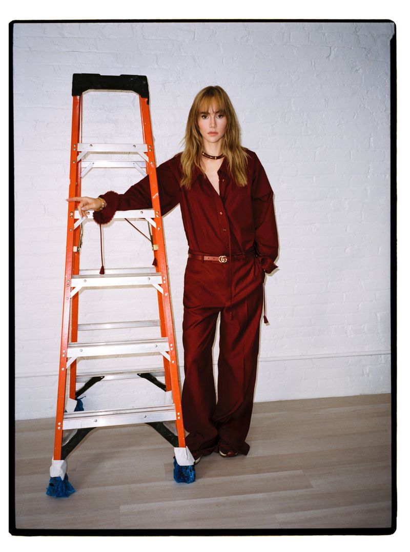 A woman in a burgundy jumpsuit standing next to an orange and aluminum ladder in a photo studio with a white brick background.
