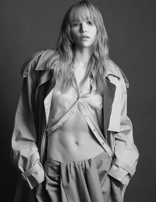 Suki Waterhouse wearing an open trench coat and button-down shirt against a dark studio background in a black and white portrait.