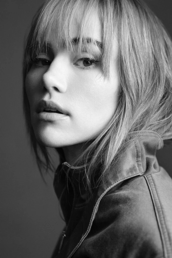 Close-up black and white portrait of a woman with bangs and a jacket looking directly at the camera.