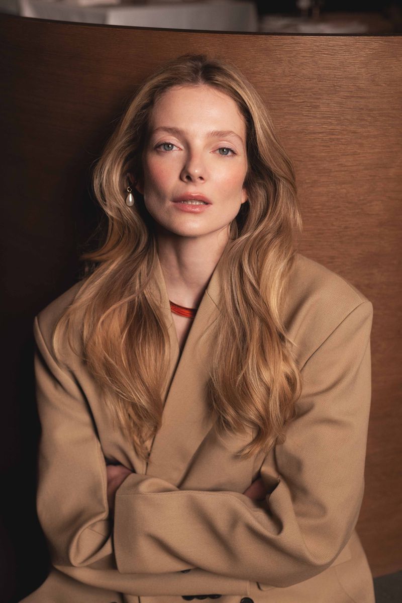 Portrait of a blonde woman with arms crossed wearing a tan blazer against a paneled background in a close-up shot with soft lighting.