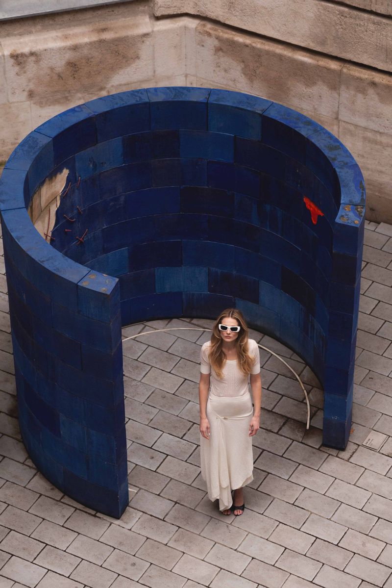 Woman in flowing beige dress and sunglasses standing in a courtyard near blue architectural structure on a patterned brick surface.