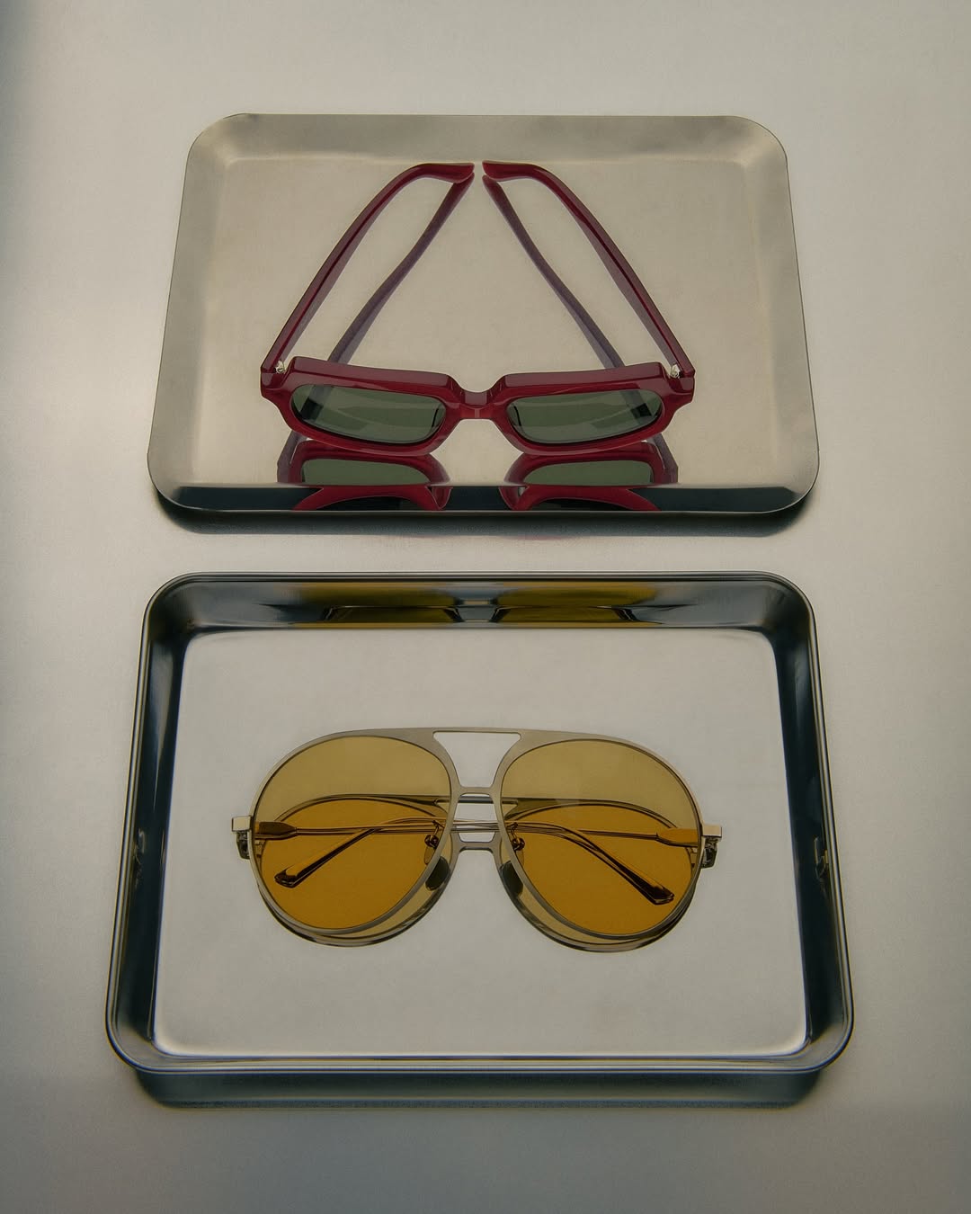 Two pairs of sunglasses, one red rectangular pair and one yellow aviator pair, are presented on silver trays against a plain background.