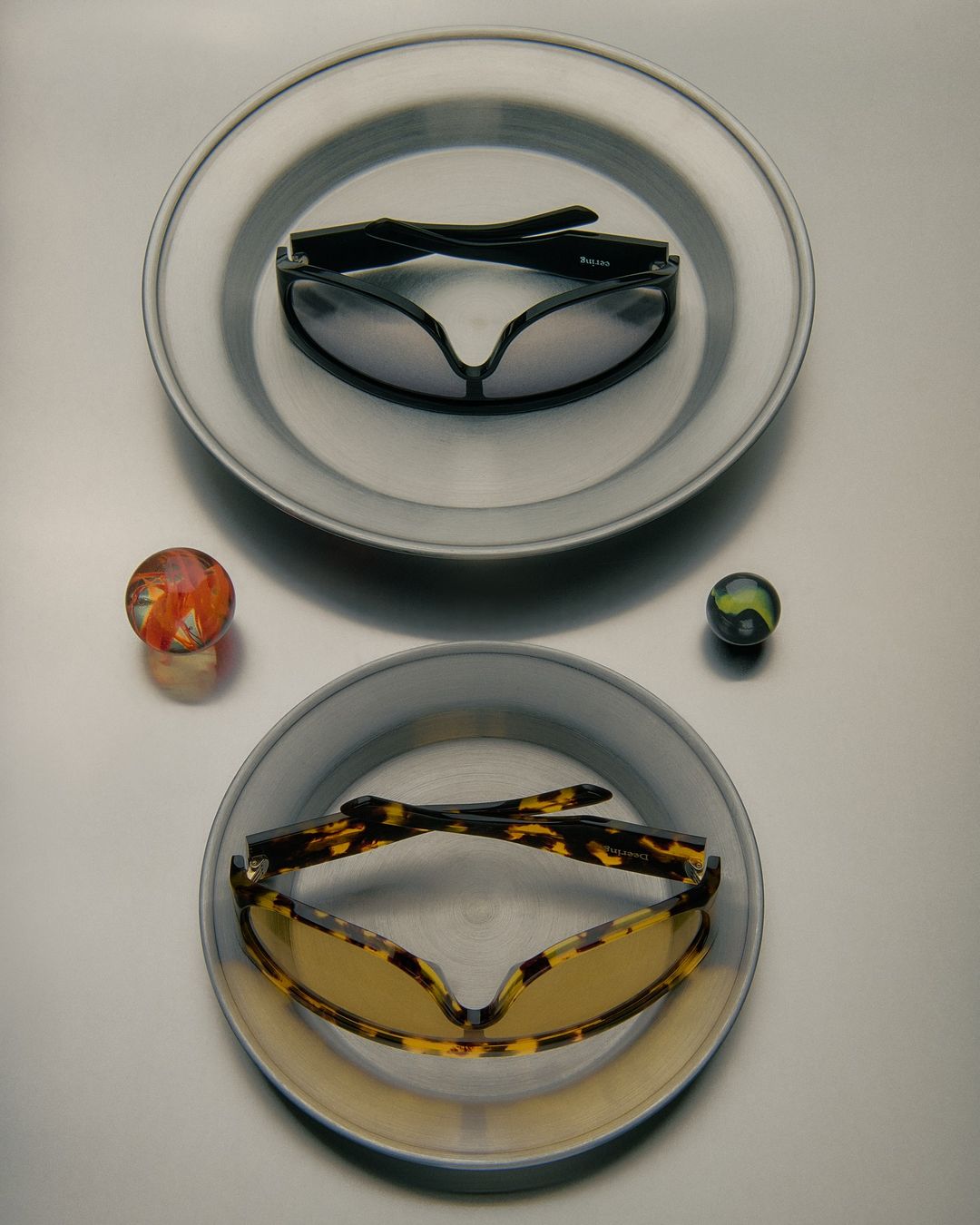 Two pairs of sunglasses sit on silver plates with glass marbles, set against a light gray background in a studio setting.