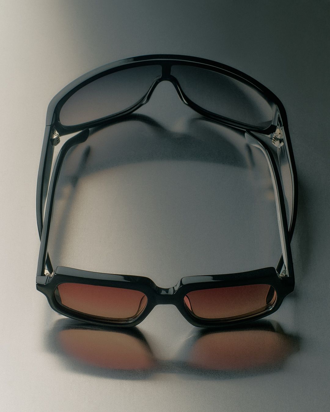 Two pairs of sunglasses are stacked on top of each other, each with a different color gradient on the lenses, against a grey background.
