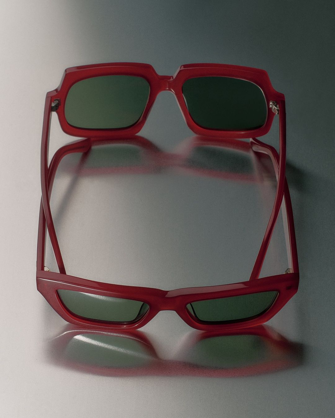 Red sunglasses with green lenses are reflected on a shiny gray surface. The image showcases eyewear fashion with a simple, clean background.