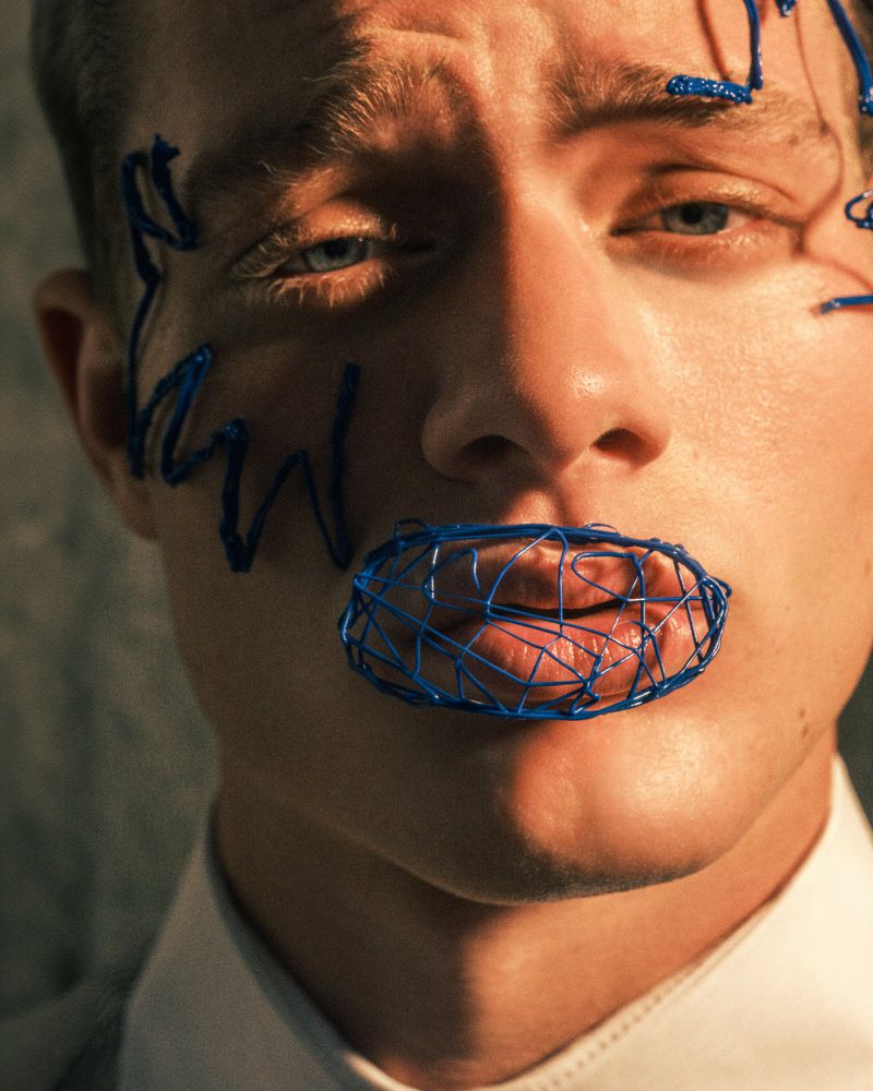 Close-up portrait of a light-skinned adult male with blue line art on his face and lips, wearing a white collared shirt.