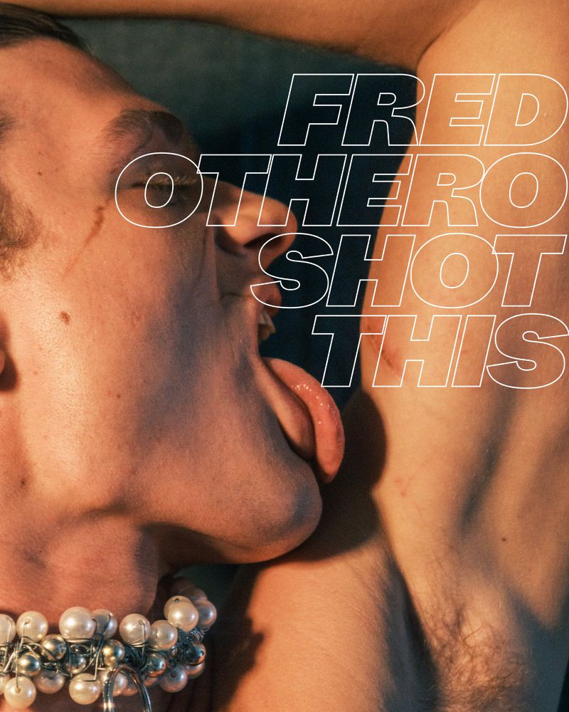 Close-up of two adult men's faces, one with an extended tongue touching the other's skin, adorned with a pearl beaded necklace. 