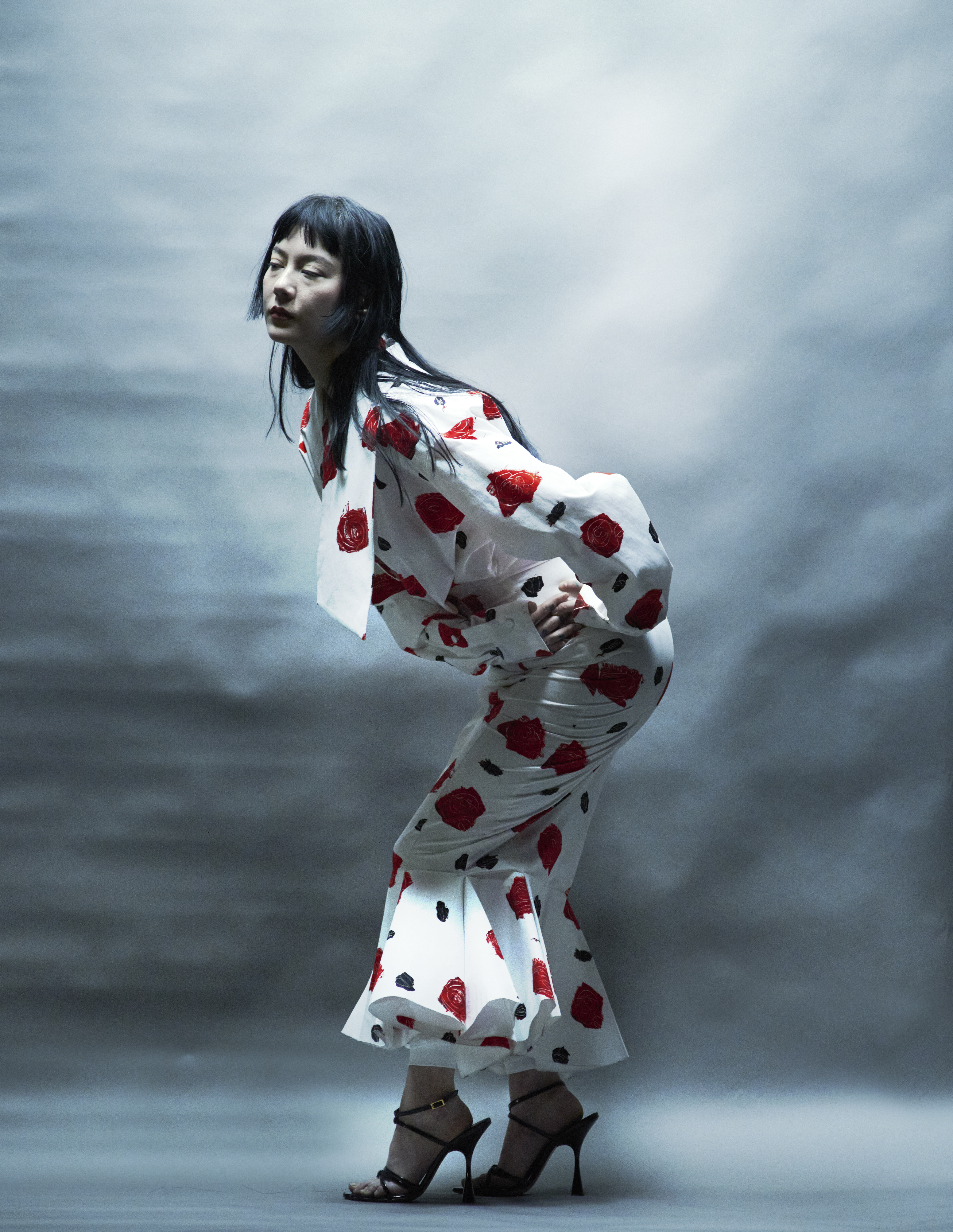 Fashion shot of an Asian woman in a white and red floral patterned outfit bending over with hands on her hips in a studio setting.