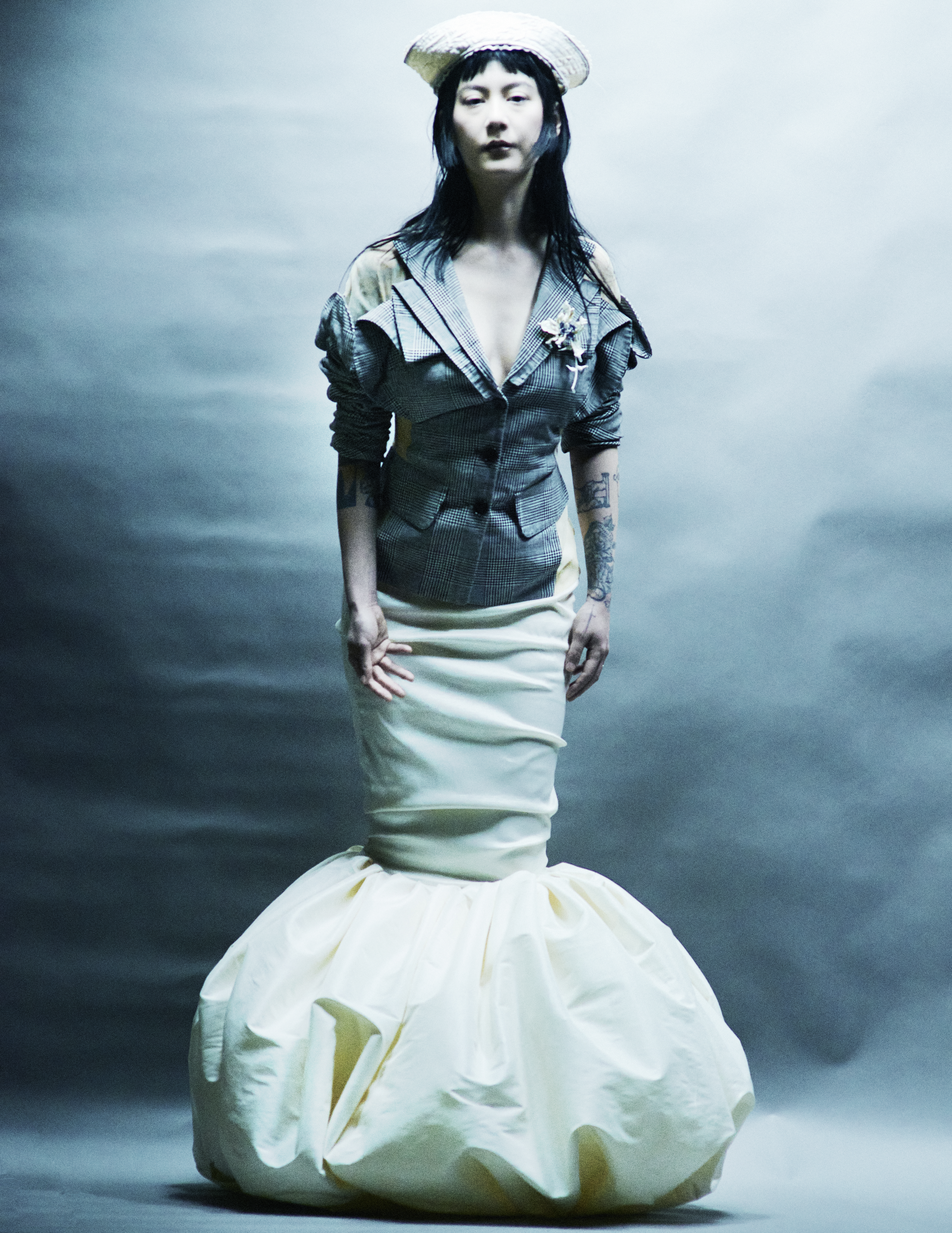 An Asian woman posing in a grey jacket with a decollete and a white balloon skirt, standing in front of a grey cloudy background.