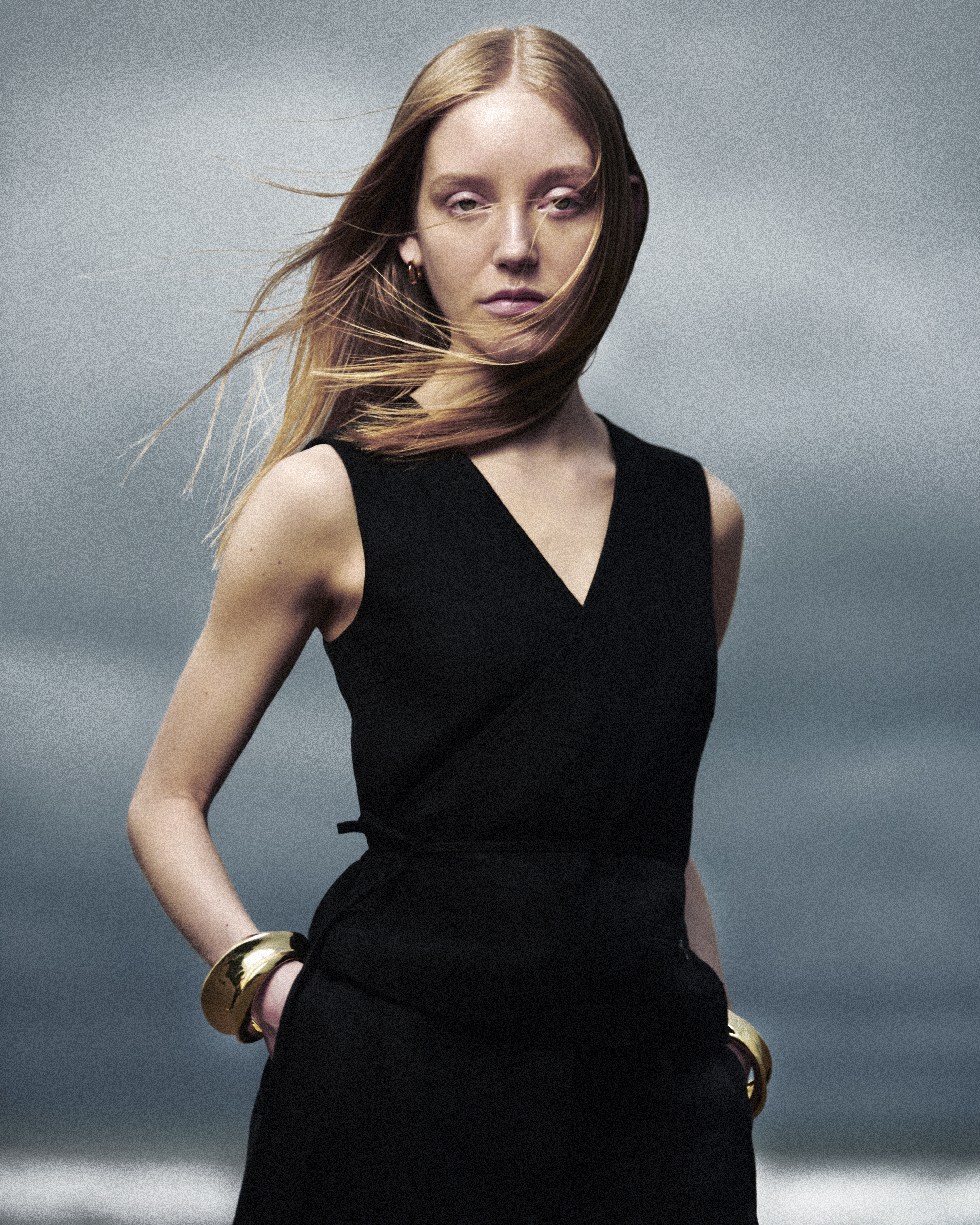 Portrait of a blonde woman in a black dress with windblown hair against a cloudy sky background, showcasing fashion and beauty.