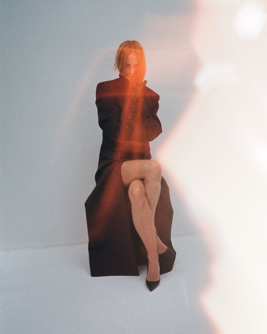 A solo adult woman sits in a photo studio, elegantly dressed in a dark coat and heels, bathed in bright light with lens flares.