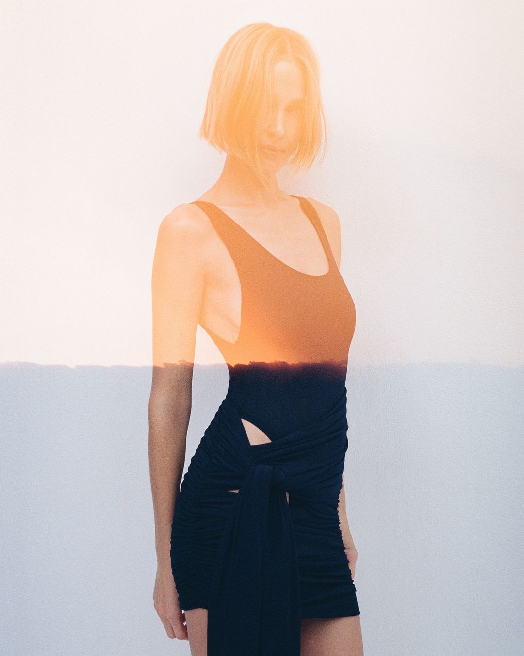 Blonde woman poses in black swimsuit with a sunset double exposure for a fashion editorial image against a white background.