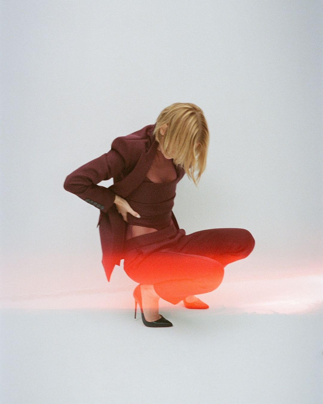 A fashion model crouches in a photo studio wearing a matching red blazer, wrap top, and pants with black high heels against a white backdrop.