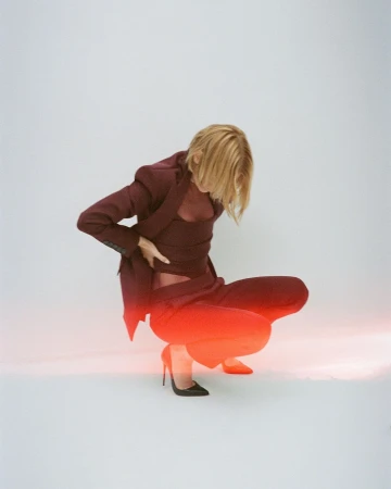 A fashion model crouches in a photo studio wearing a matching red blazer, wrap top, and pants with black high heels against a white backdrop.