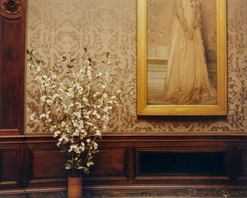 Interior view of a room with damask wallpaper, ornate wood panelling, a framed portrait, and a vase of white flowering branches.