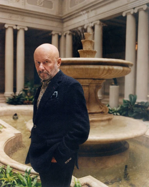 Portrait of a mature man in a dark blazer, posed in a colonnaded courtyard with a tiered fountain, plants, and architectural details.