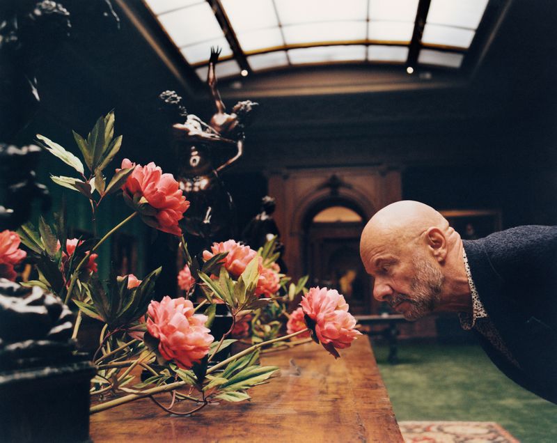 Elderly man in black jacket looks at pink peonies in elaborate display with dark statue in background, bright skylight above.