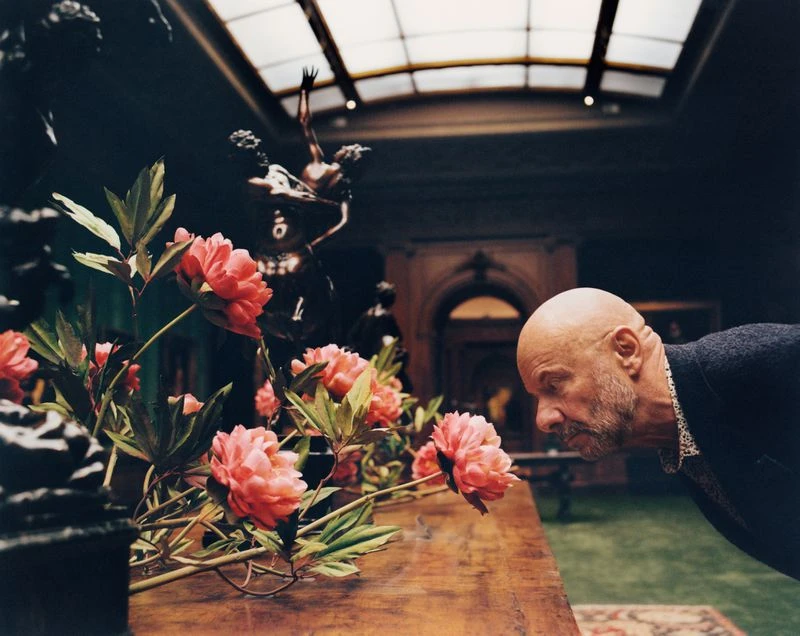Elderly man in black jacket looks at pink peonies in elaborate display with dark statue in background, bright skylight above.