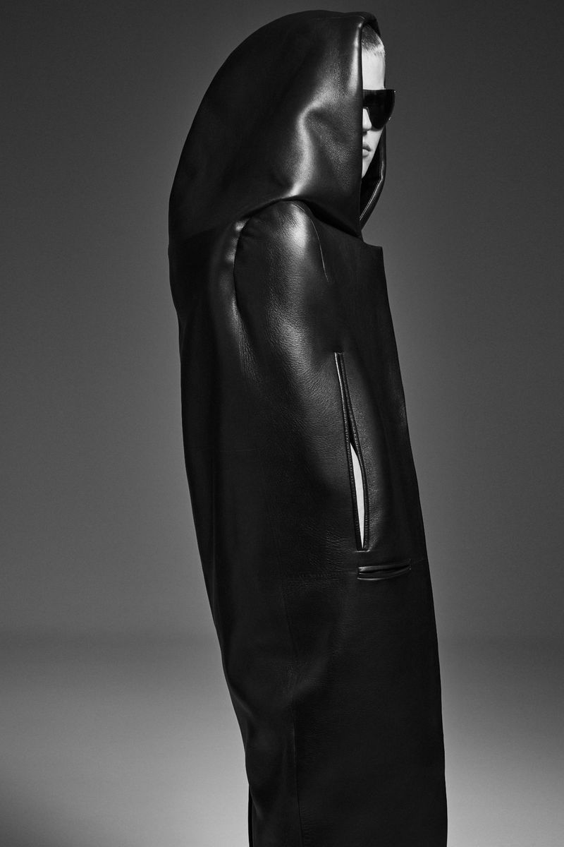 Monochrome studio shot of a person wearing a black hooded, leather-like coat and sunglasses, set against a graduated gray background.