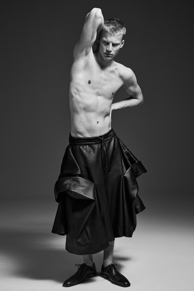 Monochromatic portrait of a shirtless male model wearing a dramatic leather skirt and black dress shoes in a studio setting.
