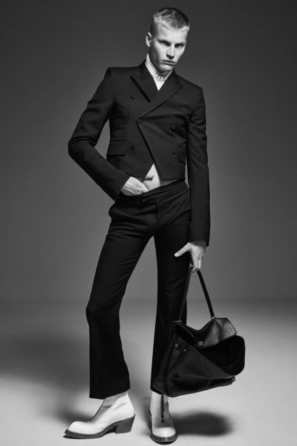 Black and white studio shot of a slender male model wearing a cropped suit jacket and holding a bag. He is wearing white boots.