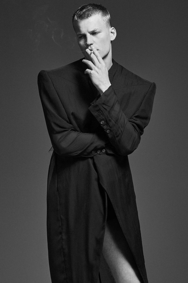 A black and white portrait of an adult man smoking a cigarette, wearing a dark coat, standing in a photo studio.