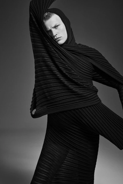 Monochrome studio portrait of a fair-skinned adult male wearing a draped hooded outfit with a serious expression, against a grey backdrop.