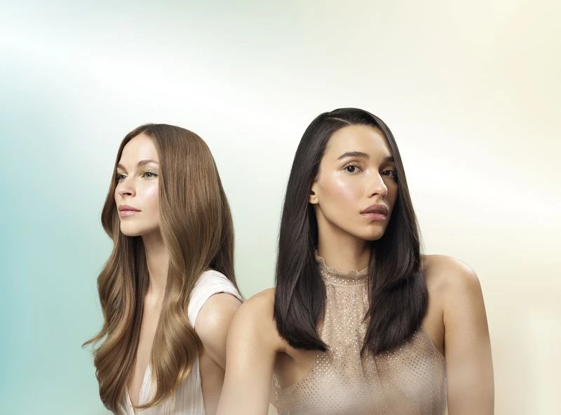 Two women pose in a beauty shot, showcasing dark brunette and light brown wavy hairstyles, makeup, and formal dresses on a soft gradient backdrop.