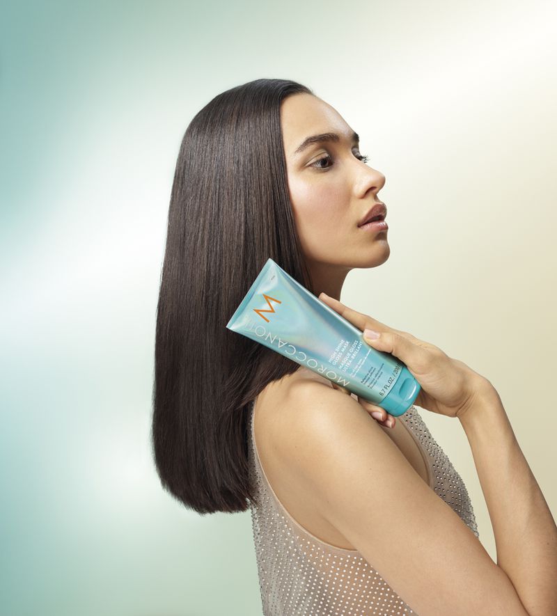 Beauty shot of a woman with straight dark hair holding a teal Moroccanoil product tube against a soft, graduated yellow-to-blue background.