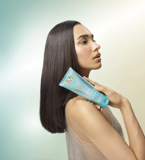Beauty shot of a woman with straight dark hair holding a teal Moroccanoil product tube against a soft, graduated yellow-to-blue background.