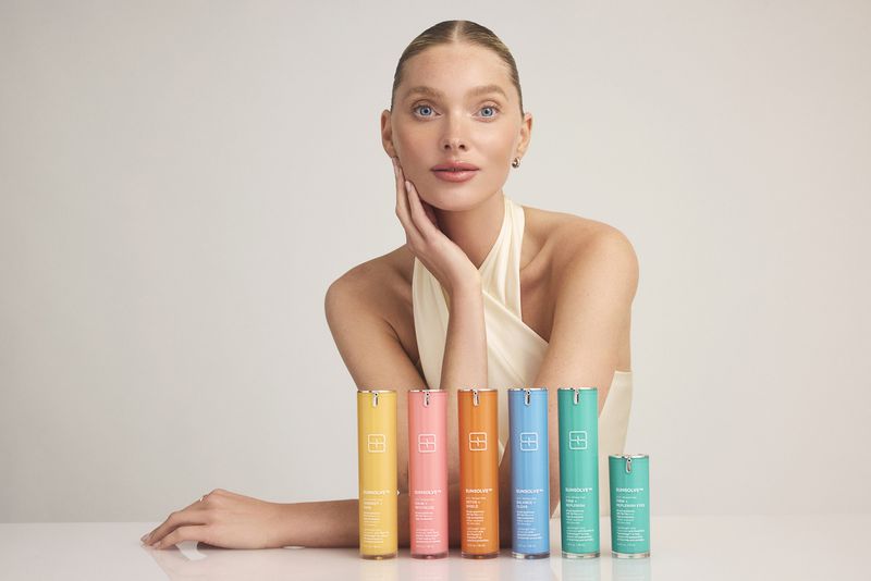 Blonde woman poses with a line of colorful skincare product bottles on a white table against a light background in a studio setting.