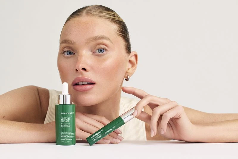 Blonde woman with blue eyes posing with Sunsolve green and silver skincare products on a white surface in a beauty product shot.