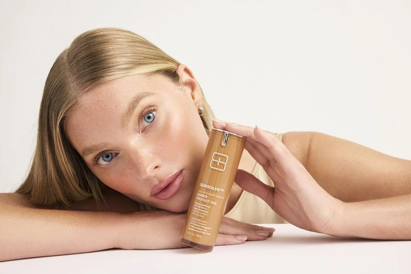 Blonde woman with blue eyes poses with skincare container in her hands against white backdrop in an eye-level, medium shot.