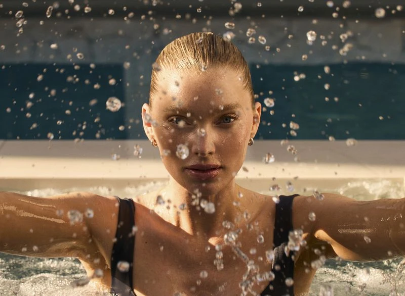Blonde woman stands in a pool with splashing water. Beauty shot with a close-up framing, showcasing clear skin and gold earrings.