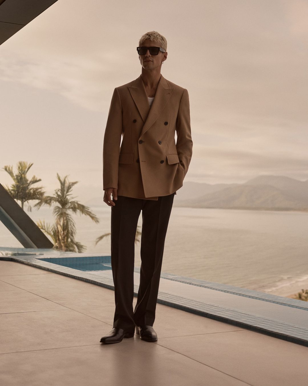Tan-suited man with sunglasses stands near pool overlooking ocean landscape, showcasing fashion and scenic views on a sunny day.