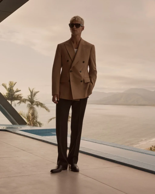 Tan-suited man with sunglasses stands near pool overlooking ocean landscape, showcasing fashion and scenic views on a sunny day.