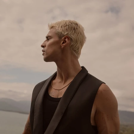 Profile portrait of a young man with bleached blonde hair wearing a black sleeveless vest against an overcast sky and water backdrop.