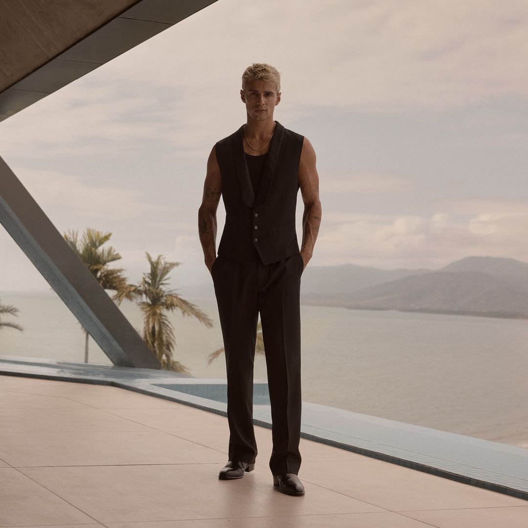 Attractive adult male in a vest and black suit stands in front of tropical ocean and palm tree landscape. Fashion editorial.