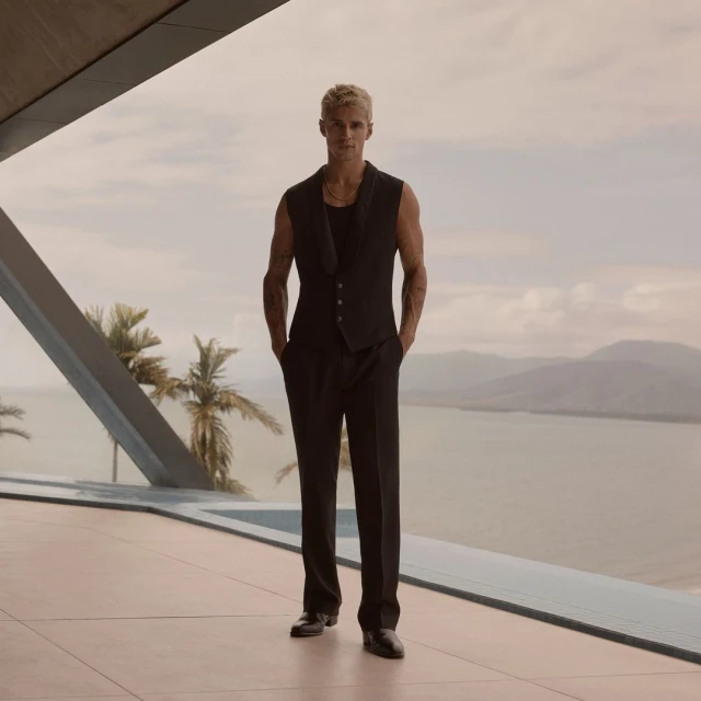 Attractive adult male in a vest and black suit stands in front of tropical ocean and palm tree landscape. Fashion editorial.